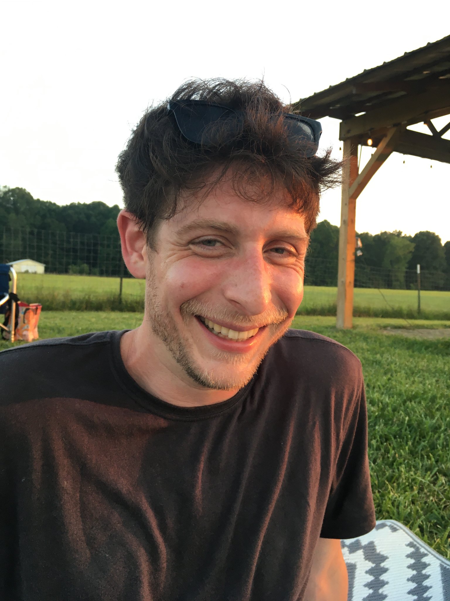 An Indigenous person with brown hair and a light beard and mustache smiles for the camera. He is sitting in front of a grassy field with a forest backdrop under an open sky and wearing a black t-shirt and sunglasses.
