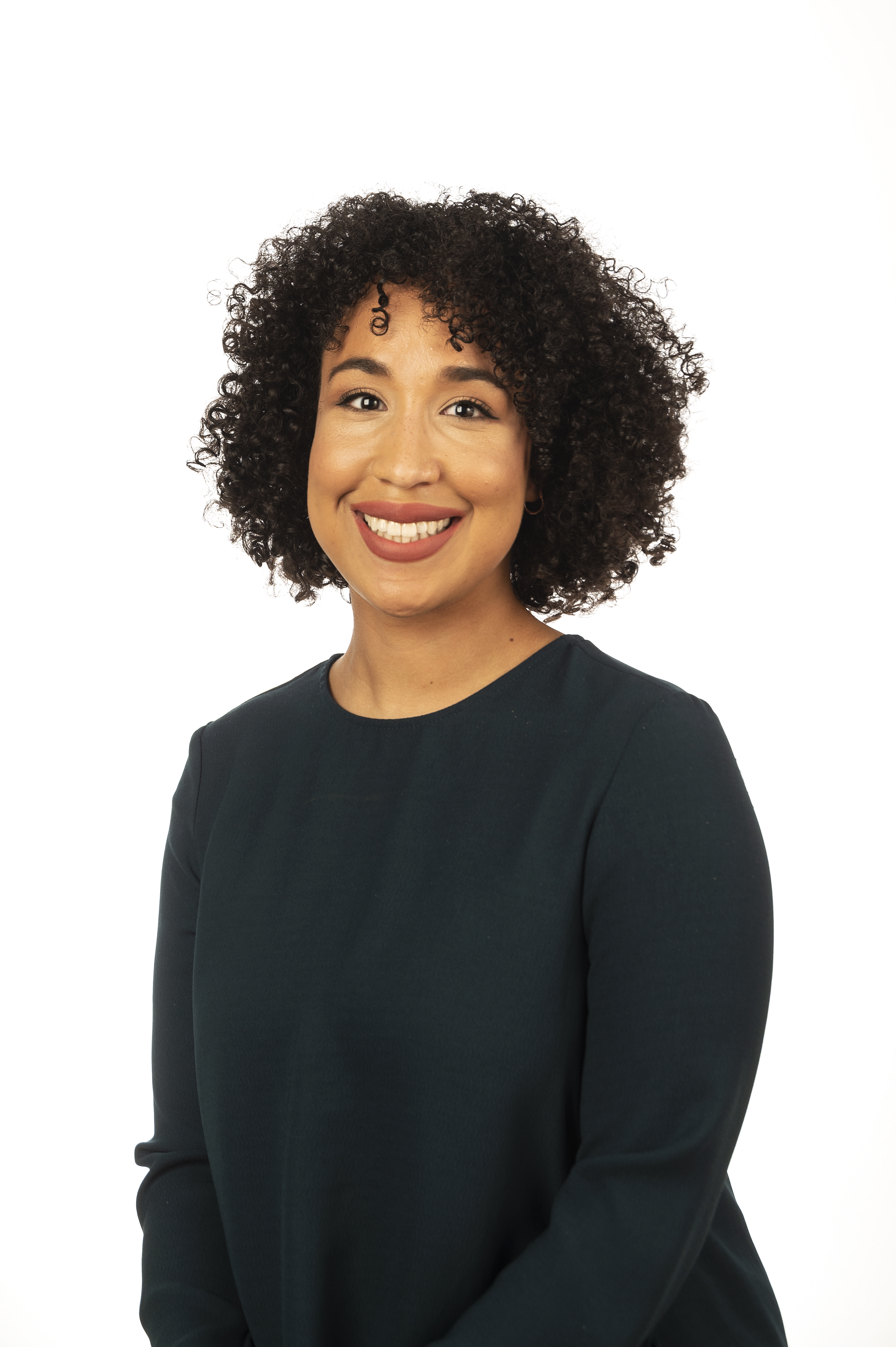 A brown woman looks at the camera in front of a white background. She has curly black hair and brown eyes and is wearing a long-sleeved black shirt.