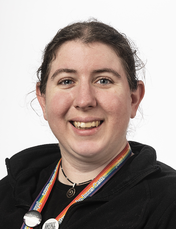 A white nonbinary person looks at the camera in front of a white background. She has brown hair and blue eyes and is wearing a black shirt with a rainbow lanyard.